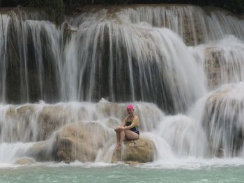 Woman sitting by waterfall on rock