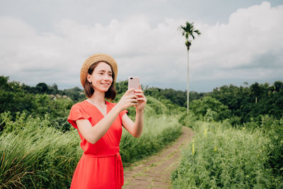 Portrait of smiling young woman holding camera while standing by plants against sky