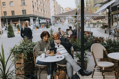 Senior woman sharing smart phone with elderly male friend having coffee at sidewalk cafe