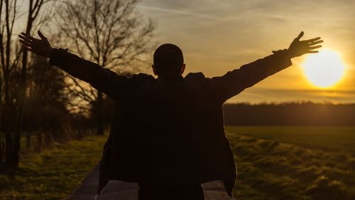 Rear view of silhouette man with arms outstretched standing on field against sky during sunset