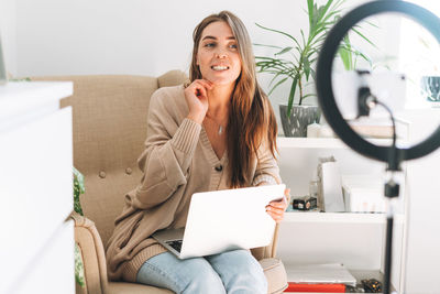 Young woman blogger records video on mobile phone using ring lamp sitting on chair with laptop 