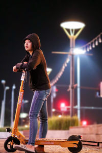 Woman standing with push scooter on footpath in illuminated city at night