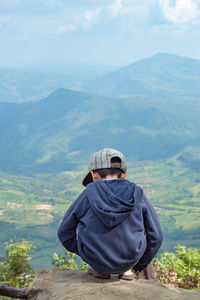 Rear view of boy sitting on cliff against landscape