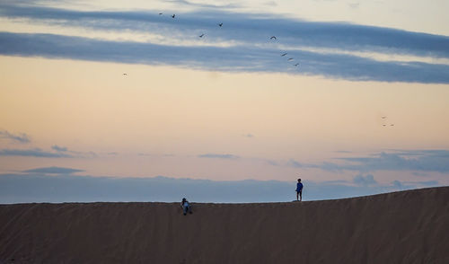 Rear view of man standing on beach against sky during sunset