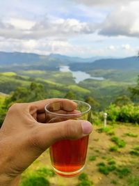 A person holding a glass of black tea against a scenic view of nature