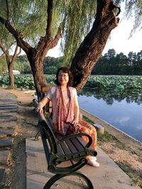 Portrait of woman sitting on bench by lake against tree