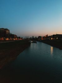 River by illuminated buildings against clear sky at sunset