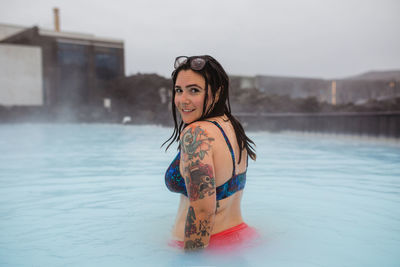 Side view of young tattooed hipster in swimsuit posing in blue water between rocks