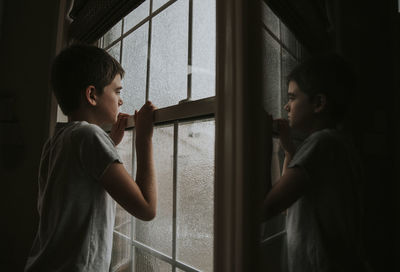 Side view of thoughtful boy looking through wet window reflecting on mirror at home