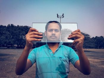 Portrait of young man holding camera against sky