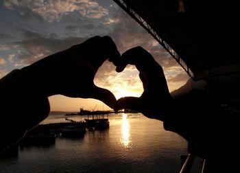 Cropped silhouette hands making heart shape by sea against sky during sunset