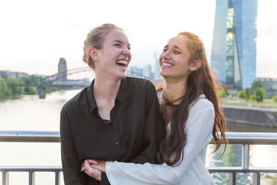 Cheerful young women embracing while standing against railing on bridge in city