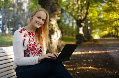 Portrait of smiling beautiful woman using laptop while sitting on bench at park