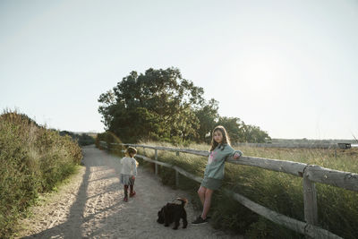 Rear view of two girl walking on field with dog against clear sky