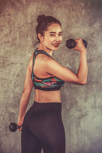 Portrait of young woman holding dumbbells while standing against wall