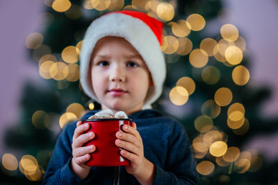 Little boy in santa hat holding a cup of hot chocolate with marshmallow