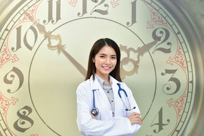 Portrait of smiling female doctor with arms crossed standing against clock