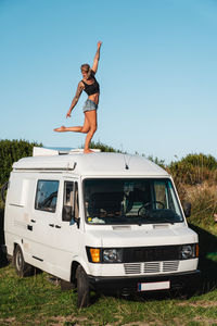 Full body of pleasant barefoot hippie female traveler standing on roof of white camping van parked in nature during road trip