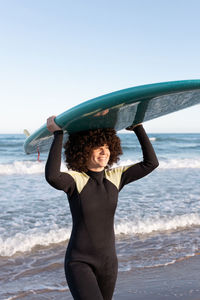 Side view of young happy female surfer in wetsuit with surfboard standing holding surfboard above head looking away on seashore washed by waving sea