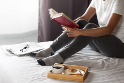 Low section of woman reading book on bed at home