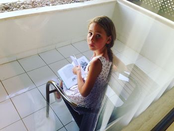 High angle portrait of girl reading book seen through glass window while sitting at home