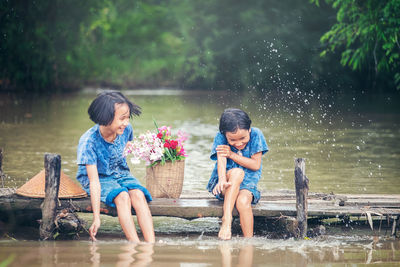 Cheerful girl splashing water on sister sitting on bridge in river