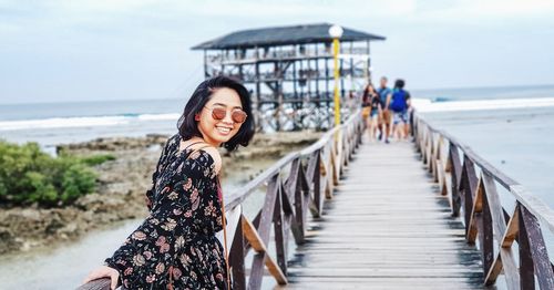 Portrait of woman wearing sunglasses while standing on pier at beach