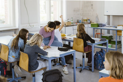 Female teacher assisting schoolgirls while sitting on desk in classroom