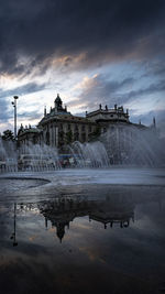 Fountain in lake by buildings against sky during sunset