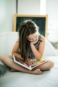 Full length of girl reading book while sitting on bed at home