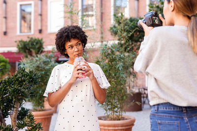 Girl photographing friend while drinking against plants