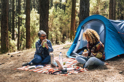 Mother and daughter having food and drink against tent in forest