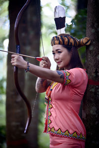 Young woman holding bow and arrow standing by tree outdoors