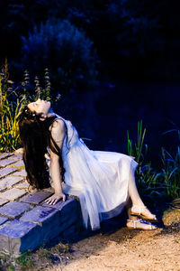 Young woman looking up while sitting by pond at night