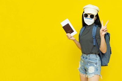 Portrait of woman wearing mask and sunglasses standing against yellow background