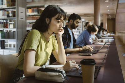 Focused young woman studying on laptop while leaning on elbow at desk in college library