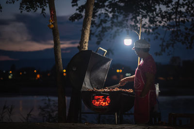 Woman cooking food on barbecue against sky at night
