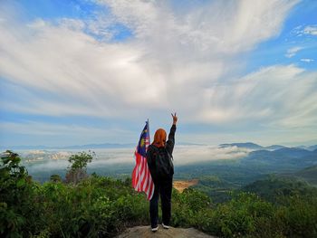 Rear view of woman standing with malaysian flag on cliff against cloudy sky