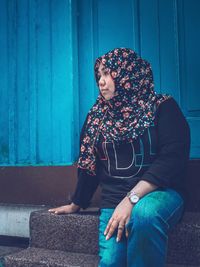 Mid adult woman wearing hijab looking away while sitting on steps against built structure