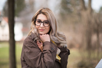 Portrait of young woman standing with hand on chin against trees