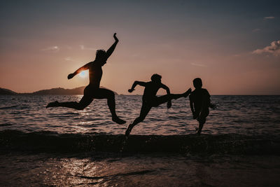 Silhouette friends jumping in sea against sky during sunset