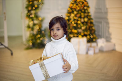 Portrait of a boy a child in a white sweater holding a gift box at the christmas tree