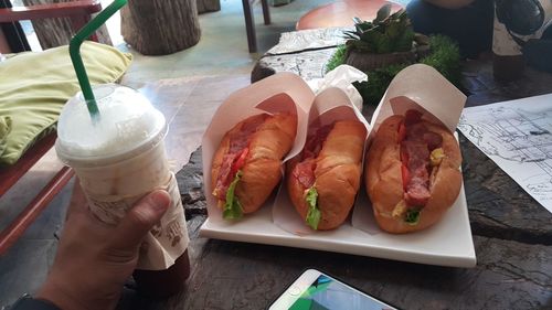 Cropped hand holding drink by hot dogs on table in cafe