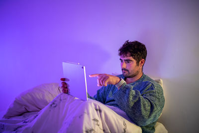 Man using digital tablet sitting on bed against wall at home