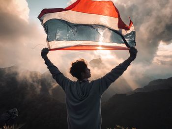 Rear view of man holding thai flag while standing against cloudy sky during sunset