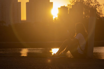 Side view of woman sitting at lakeshore during sunset