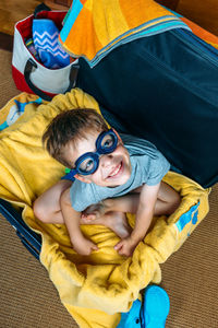 High angle portrait boy wearing swimming goggles while sitting in suitcase at home