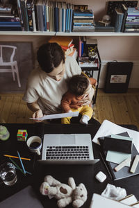 Female entrepreneur reading document while sitting with baby boy at home office