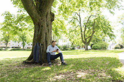 Businessman with e-scooter leaning against tree trunk at city park using smartphone