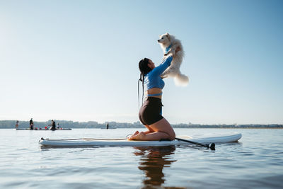 Woman raising up her dog japanese spitz while sitting on the sup board on the lake